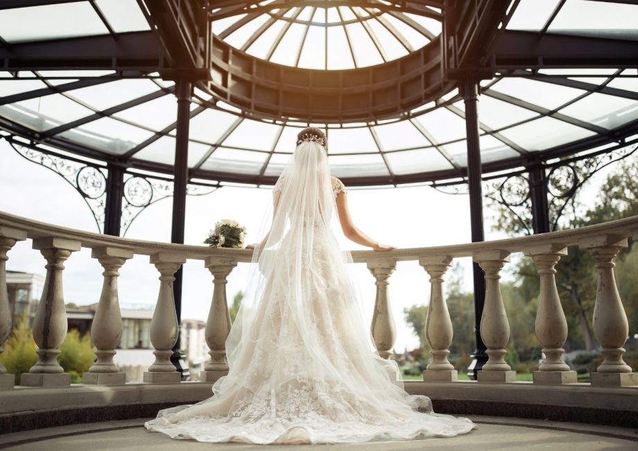 Mariée de dos en robe de dentelle ajustée à traîne et long voile, sous une rotonde