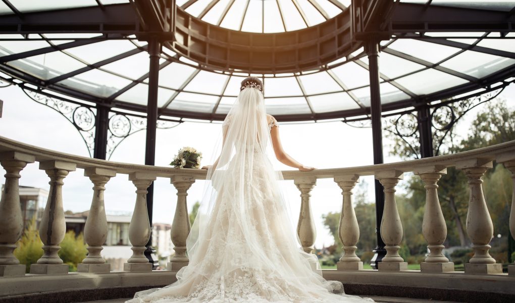 Mariée en robe blanche en dentelle avec un long voile, de dos sur un balcon baroque
