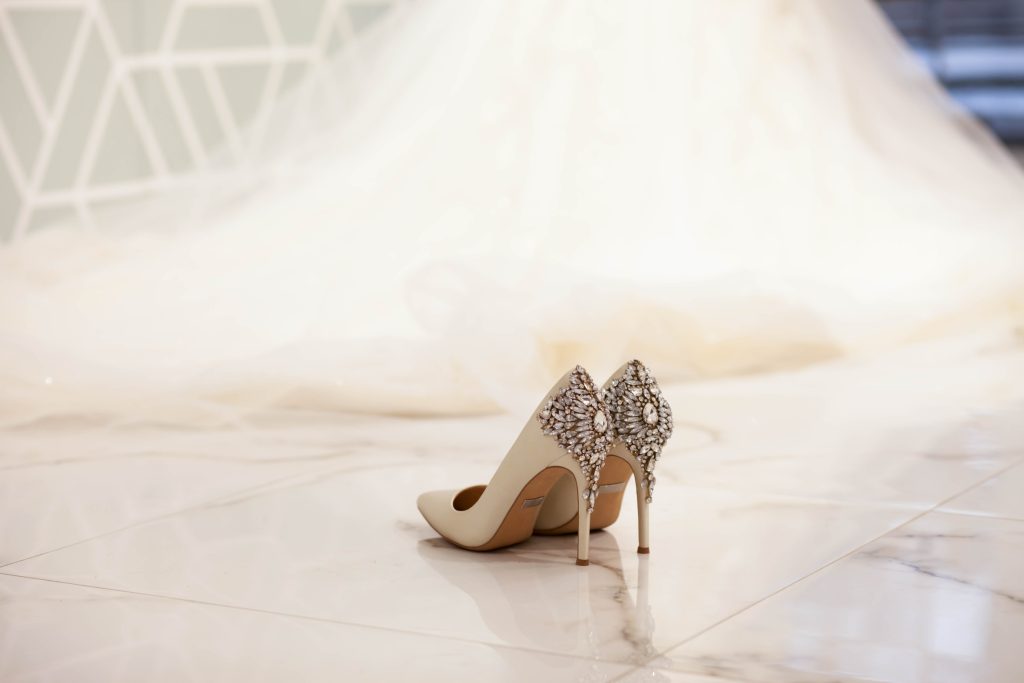 Escarpins de mariée ivoire ornés de bijoux, posés devant une longue robe de mariée